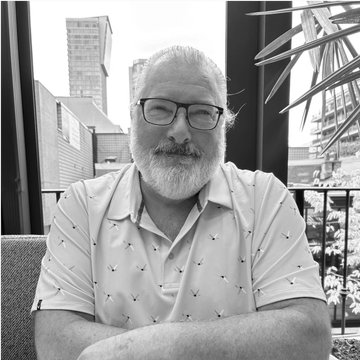 Smiling man with glasses and white beard wearing a light blue shirt, sitting outdoors.