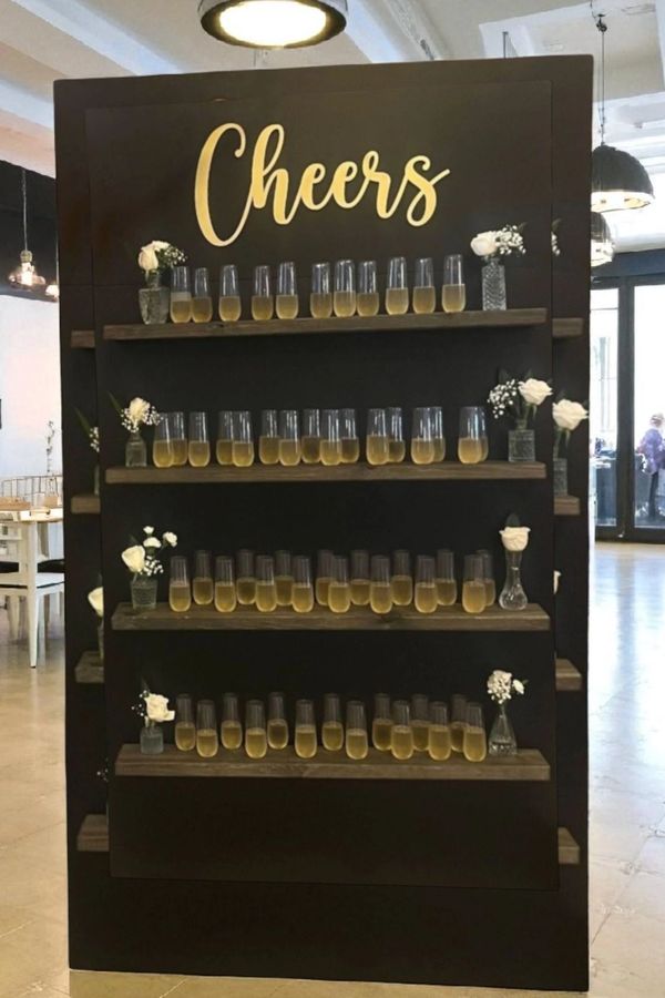 A stylish black shelf with glasses of champagne and white flowers under 'Cheers' sign.