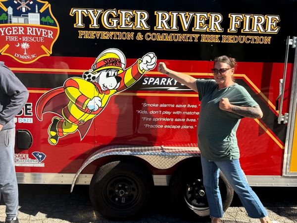 A woman poses playfully beside a fire rescue vehicle with a firefighter dog mascot.