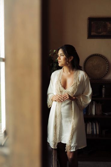 Woman in elegant white lace robe gazing thoughtfully out a window.