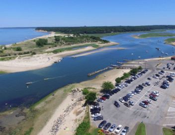 Aerial view of a coastal area with a river, beach, and parking lot filled with cars.