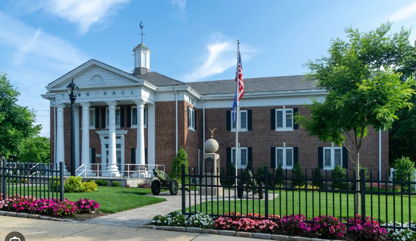 Town hall building with columns, flagpole, and well-kept garden under a clear blue sky.