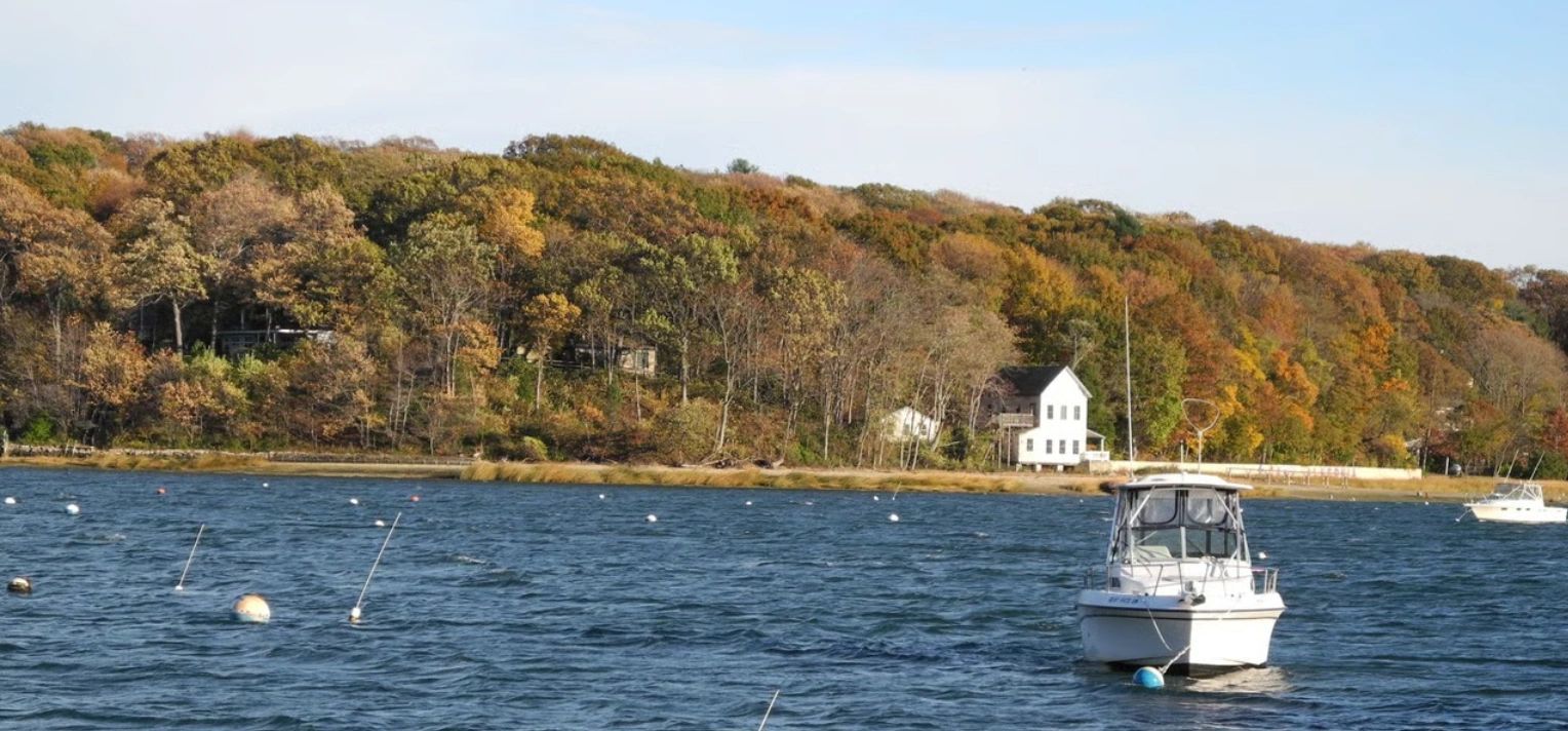 Boat anchored on blue water near autumn-colored forest and white house.