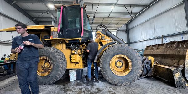 Two men working on a large yellow construction vehicle inside a garage.