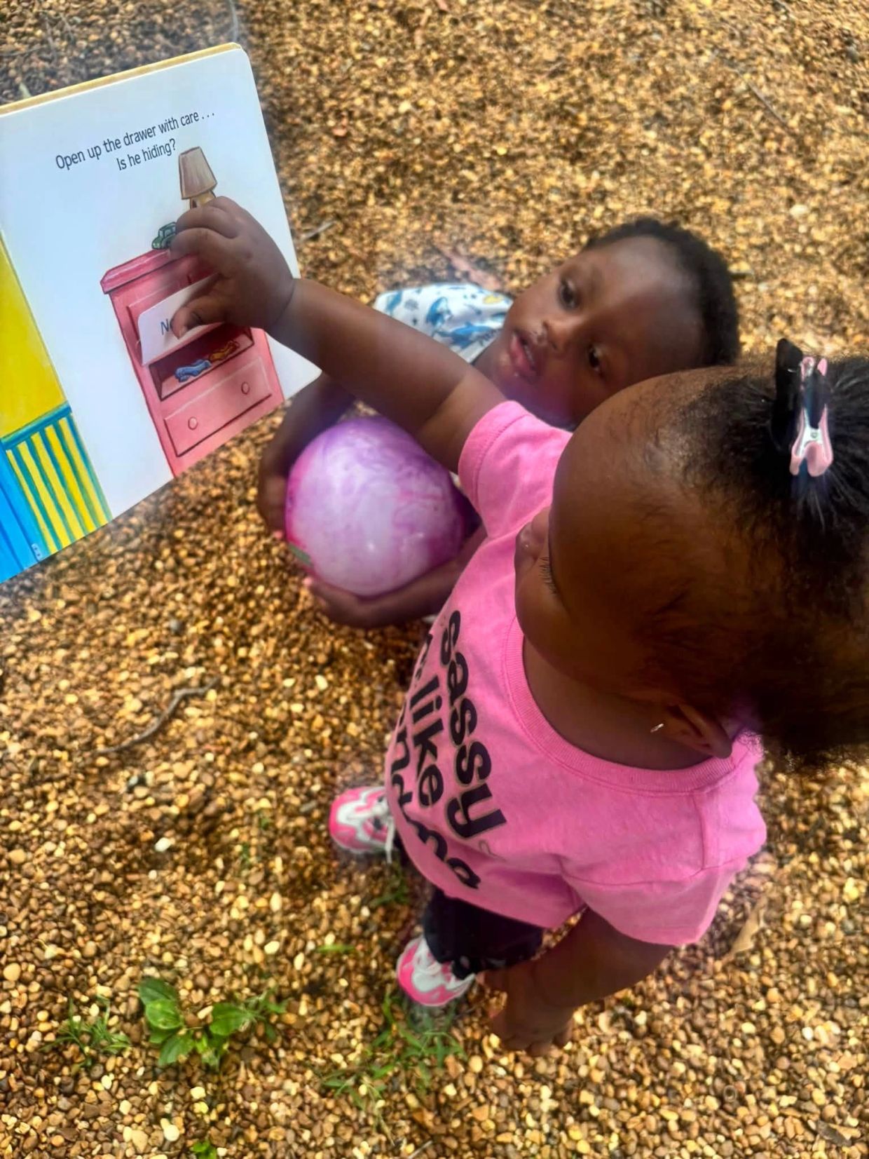 Two children reading a book outdoors, one holding a ball.