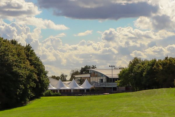 Modern building with white tent-like structures surrounded by green trees under a cloudy sky.