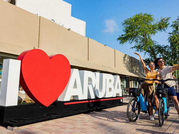 Couple happily posing on bikes beside a large I❤️ARUBA sign under clear blue skies.