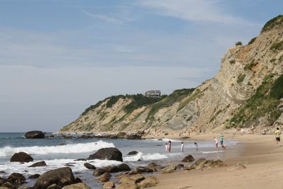 People enjoy a sunny day on a rocky beach with cliffs in the background.