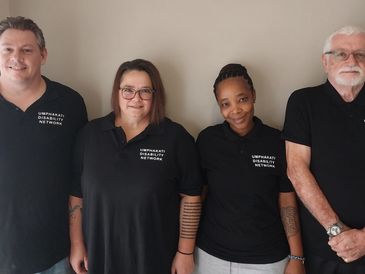 Group of four people in black shirts standing against a beige wall.