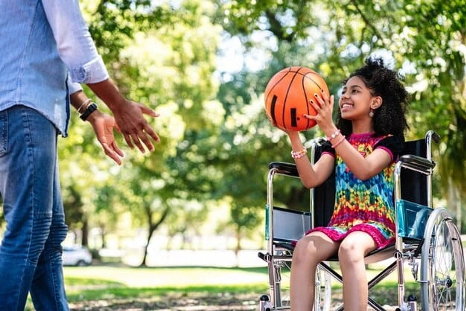A smiling girl in a wheelchair playing basketball with a man outdoors.