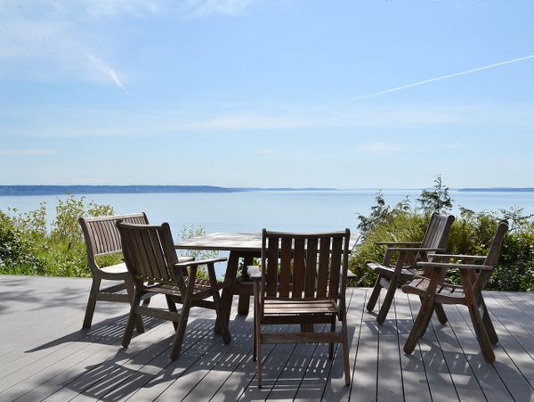 Wooden chairs and table on a deck overlooking calm water and clear sky.