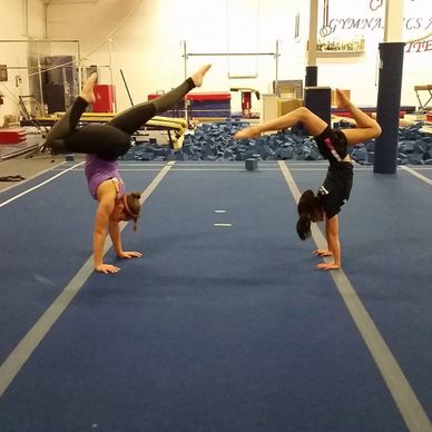 Two gymnasts performing handstands with bent legs in a gym.