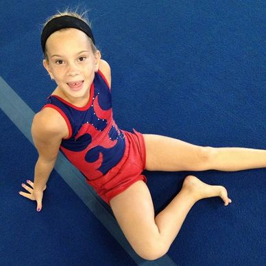 A young gymnast in red and blue leotard sitting on a blue mat.