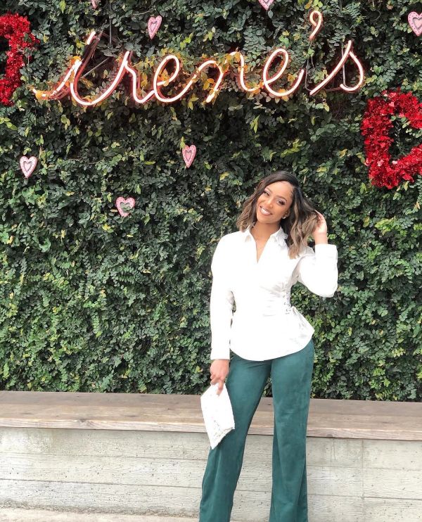Woman in white blouse and green pants posing by a leafy wall with neon sign.