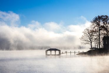 A misty lakeside scene with a small dock and bare trees under a blue sky.