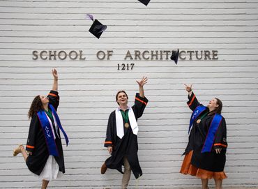 Three architecture graduates joyfully toss their caps in front of the School of Architecture.