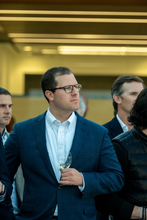 Man in a blue suit holding a glass of white wine at a social event.
