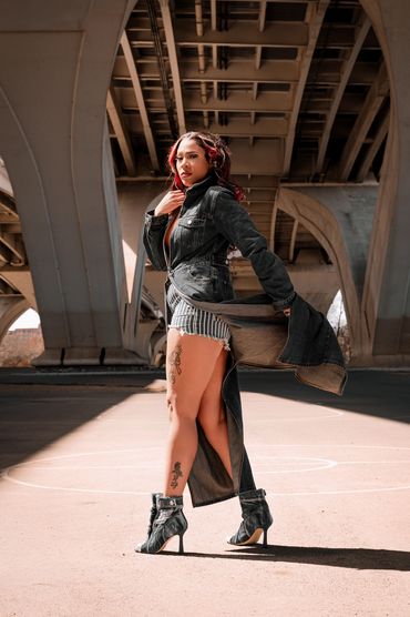 Woman posing confidently under a bridge in stylish denim outfit and high heels.