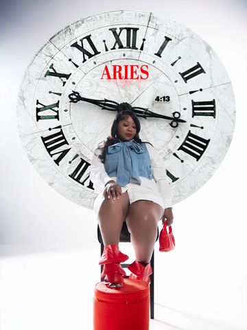 A stylish woman poses in front of a large vintage clock with Aries text.