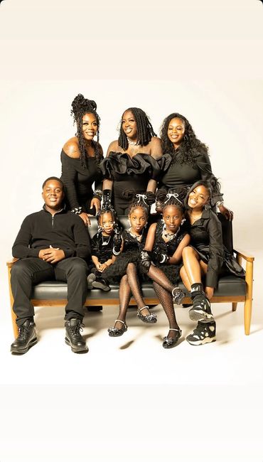 A joyful family dressed in coordinated black outfits posing on a bench against a white backdrop.