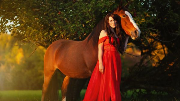 Woman in a red dress standing beside a brown horse under a tree.
