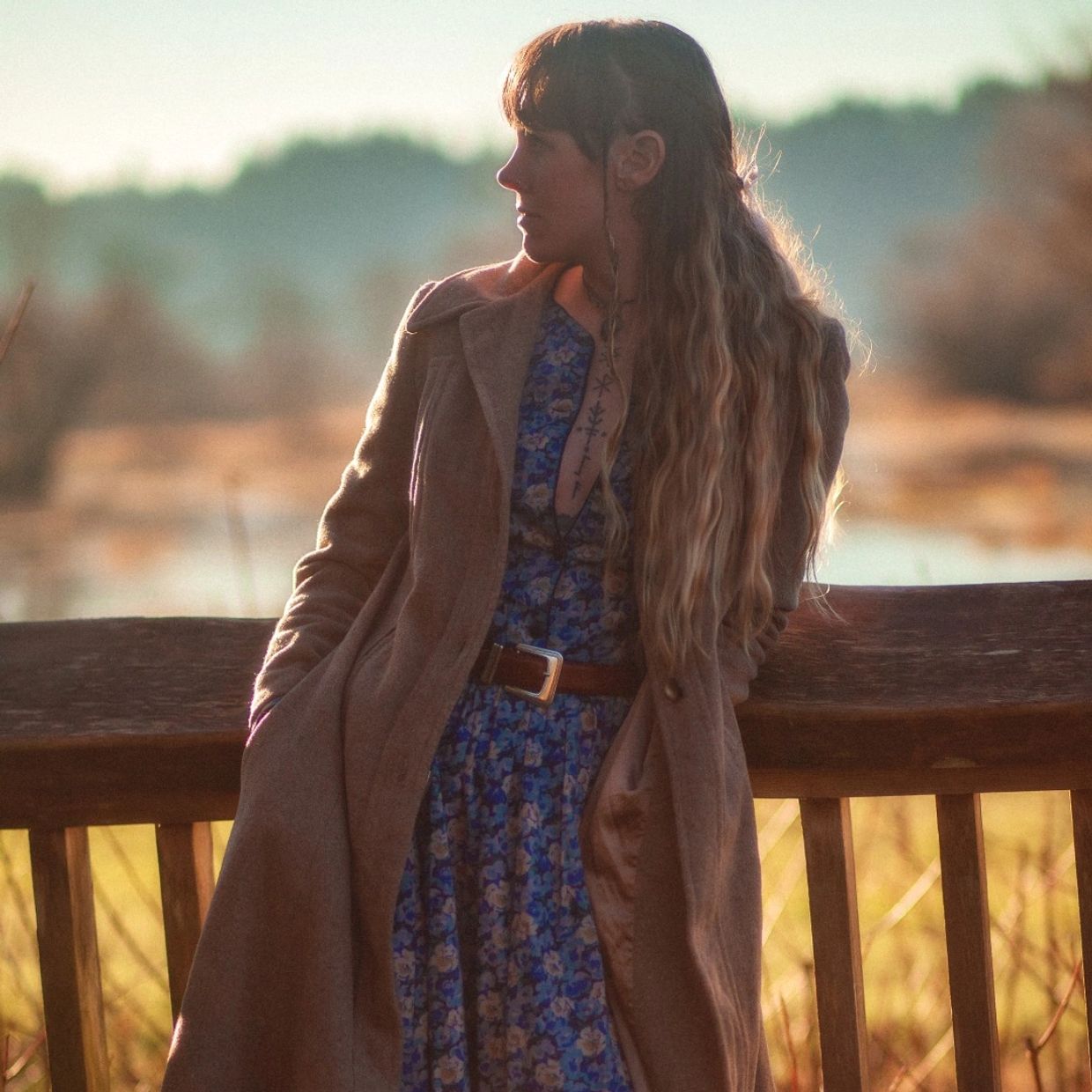 A woman in a brown coat and blue floral dress stands by a wooden bench outdoors.