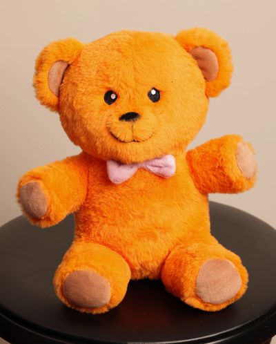 Bright orange teddy bear with a pink bow tie sitting on a black stool against a beige background.