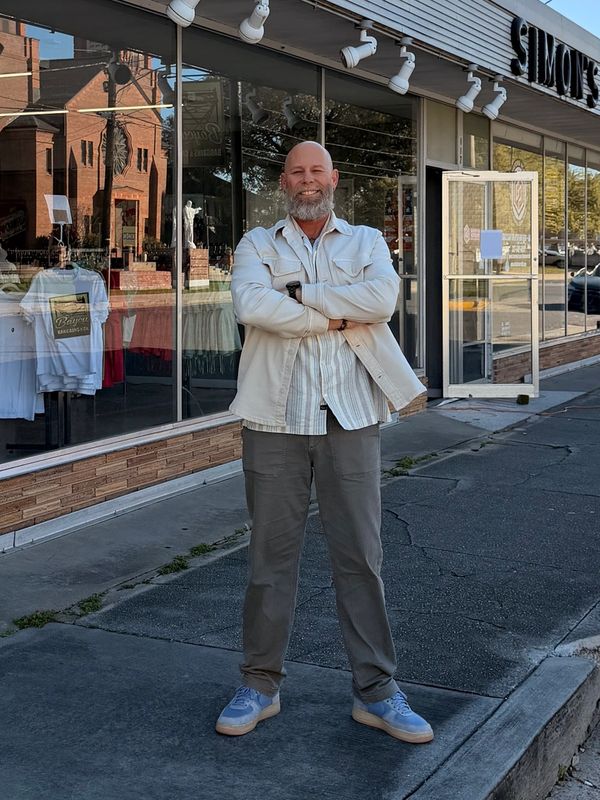 Smiling man stands outside a store called Simons on a sunny day.