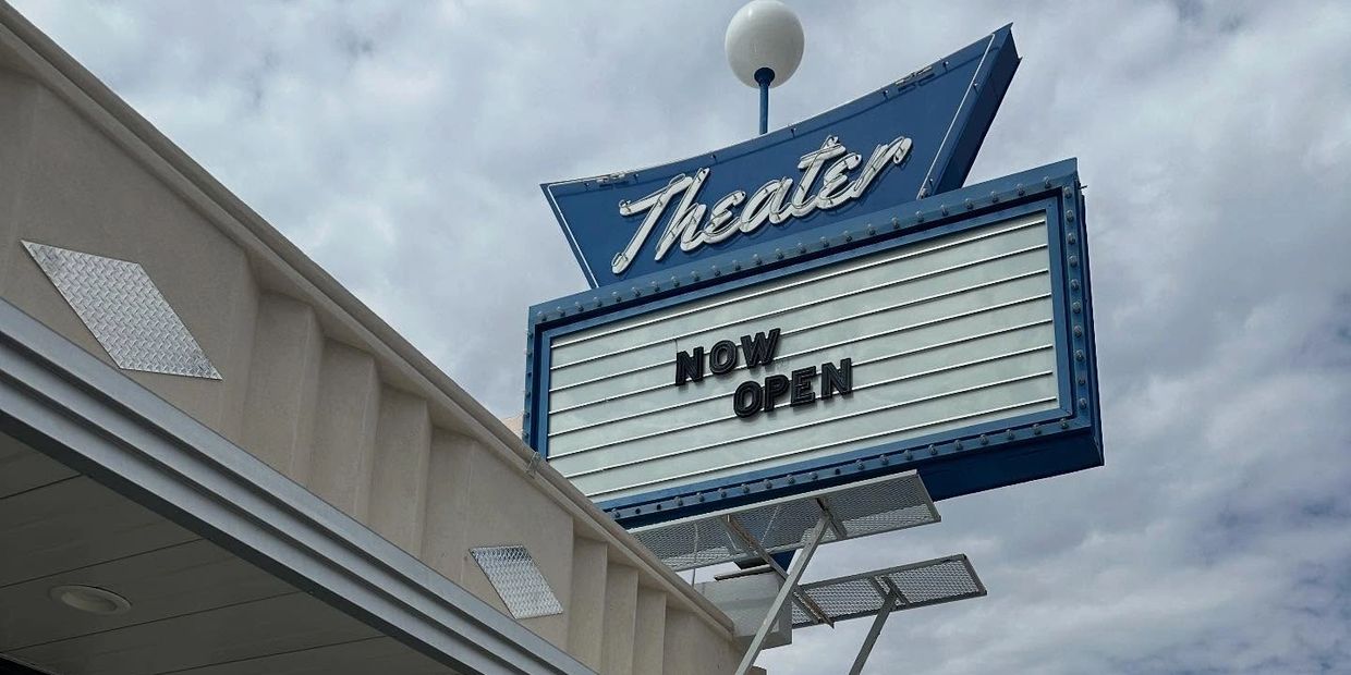 Blue theater sign reads "NOW OPEN" against a cloudy sky.