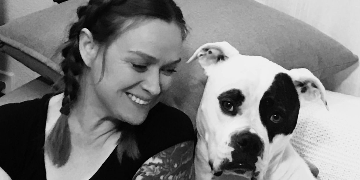 Smiling woman with braided hair sitting close to a white dog with a black patch over one eye.