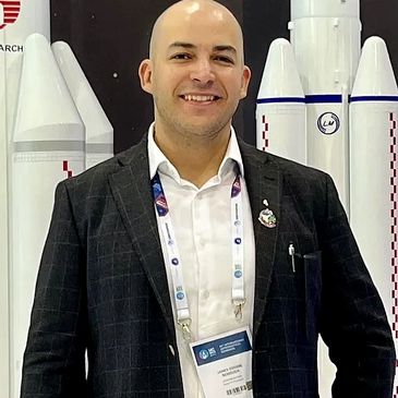 Man in a black suit smiling in front of rocket models at a conference.