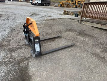 A forklift attachment with two forks on a gravel surface outdoors.