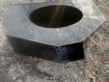 A black metal object with an oval cutout and a rectangular spout on a gravel surface.