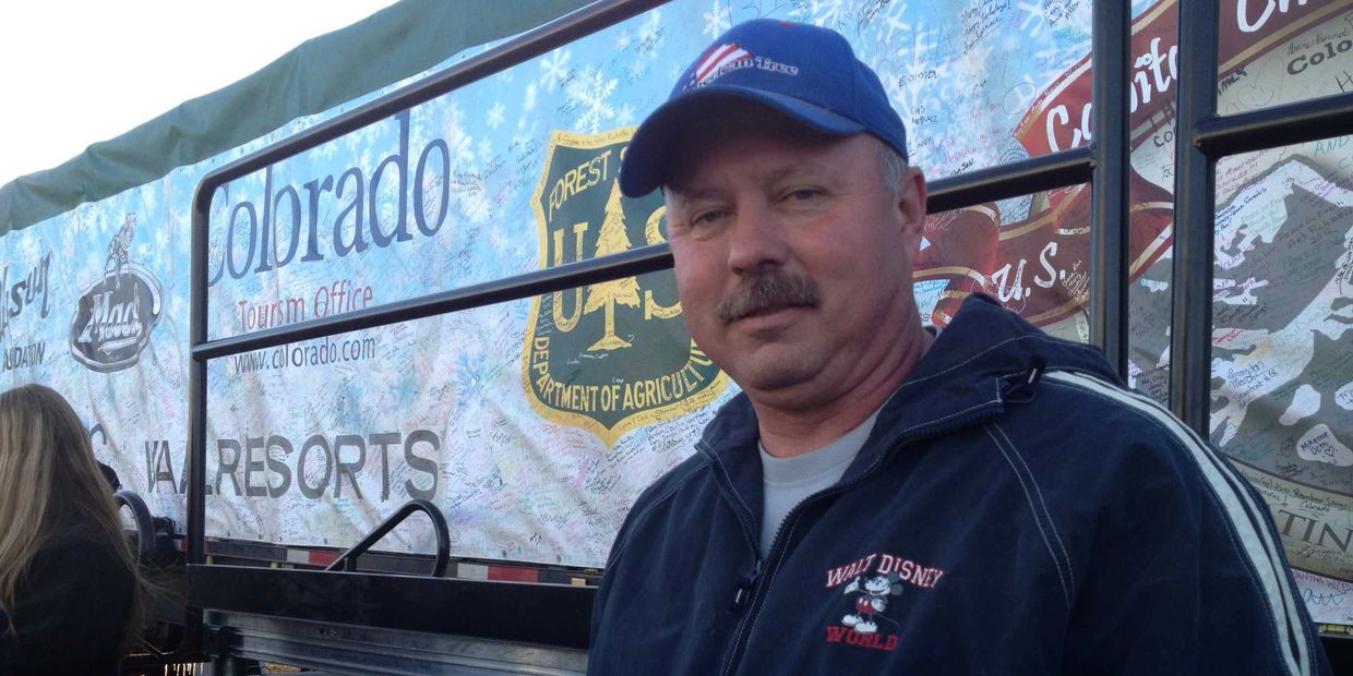 Man in a blue Walt Disney World jacket and cap stands near a Colorado tourism banner.