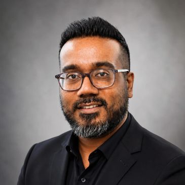 Professional portrait of a man with glasses and a beard wearing a black suit.