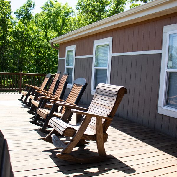 Row of wooden rocking chairs on a sunny deck beside a house.