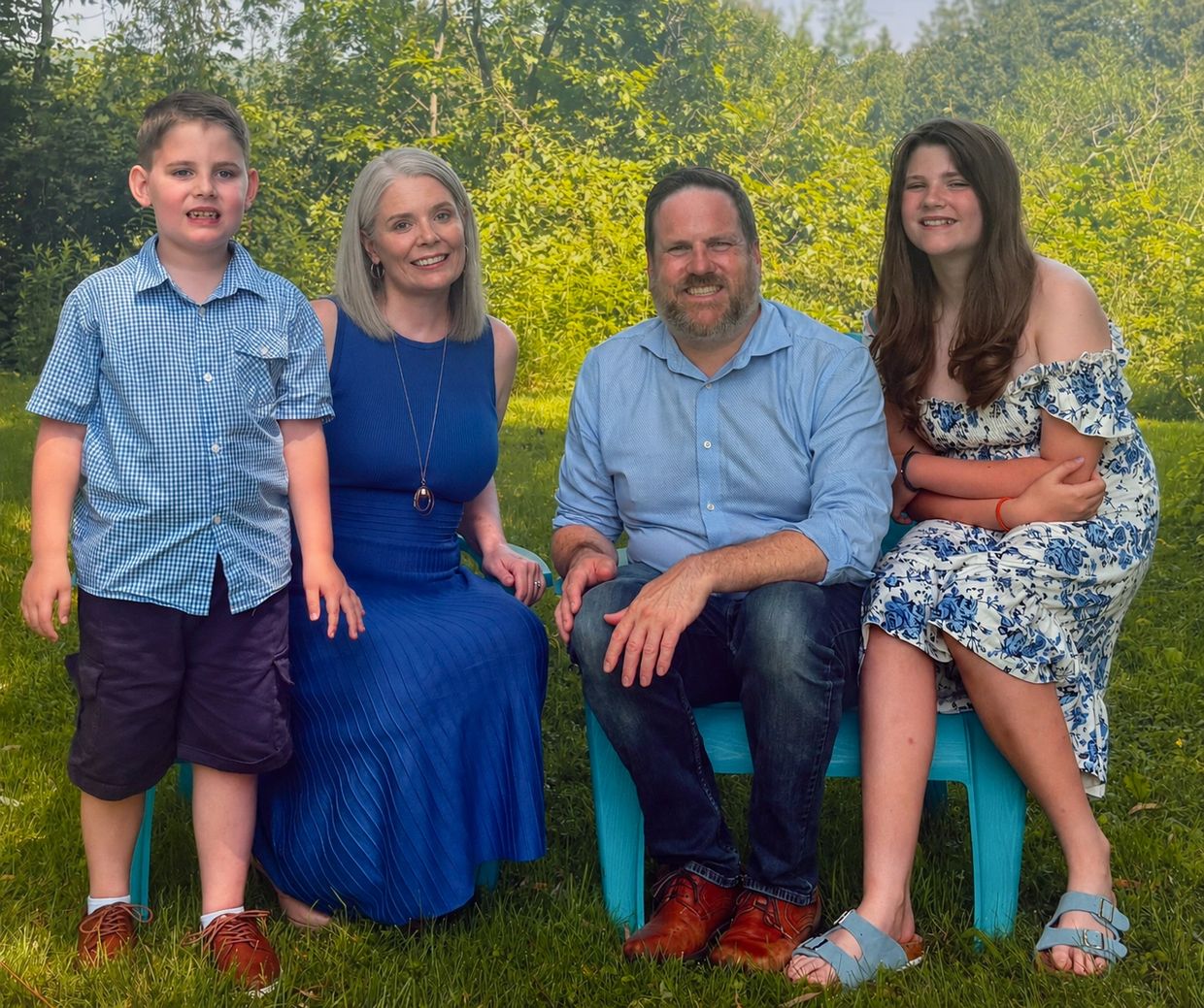 Family of four smiling outdoors in a lush green setting, with parents seated and children beside the