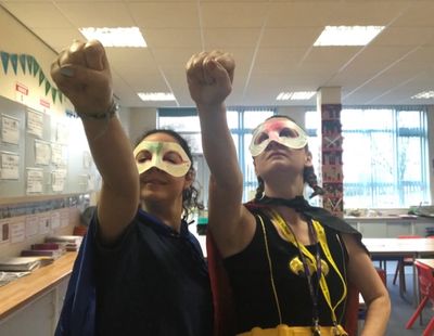 Two children in superhero masks and capes pose with raised fists in a classroom.