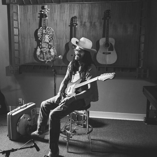Musician with guitar sitting on a stool in a room with guitars hanging on the wall.