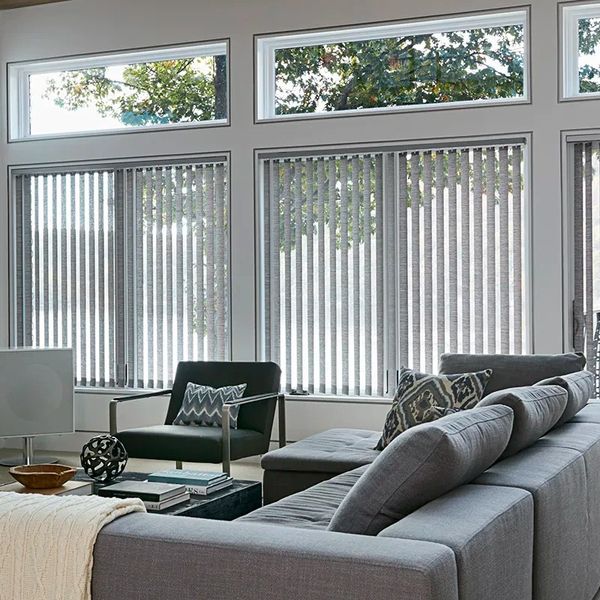 Modern grey vertical blinds installed across large floor-to-ceiling windows in a sophisticated contemporary home. The blinds feature textured fabric vanes in elegant shades of soft grey, charcoal, and stone, creating a sleek and refined aesthetic while providing effective light control and privacy. Natural daylight filters through the vertical panels, adding depth and softness to the interior while complementing large glass expanses and modern architectural lines. The space includes minimalist décor, black aluminium window frames, neutral furnishings, polished concrete or timber flooring, and subtle luxury finishes for a clean contemporary atmosphere. The non-motorized system includes discreet manual controls for practical everyday use with a streamlined appearance.

Professional installer fitting non-motorized grey vertical blinds onto large modern windows. The installation process includes accurately measuring the full window span, securely mounting the headrail track system, and evenly spacing the grey fabric vanes for a balanced and polished finish. The installer aligns the manual control mechanism, tests smooth opening and closing operation, and adjusts the rotating vanes for precise light filtering. Final scene showcases the completed installation with perfectly level grey vertical blinds enhancing the expansive windows while adding modern sophistication, privacy, and functional light control to the interior space.