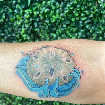 Tattoo of a sand dollar surrounded by blue ocean waves on a forearm.