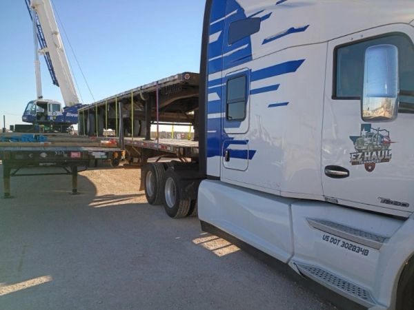 Flatbed trailer being loaded by a crane near a white truck.