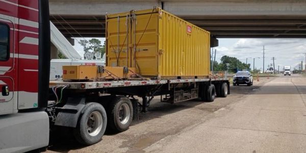 Flatbed truck carrying a yellow container parked under a bridge.