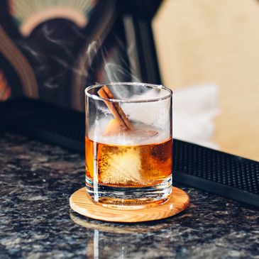 A cocktail served in a glass with a large ice cube and a cinnamon stick garnish.