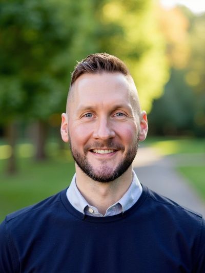 Smiling man outdoors with trees and path in background.