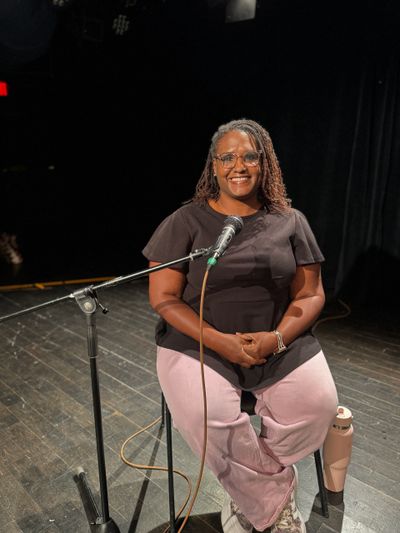 A woman smiling on stage with a microphone, sitting on a stool.