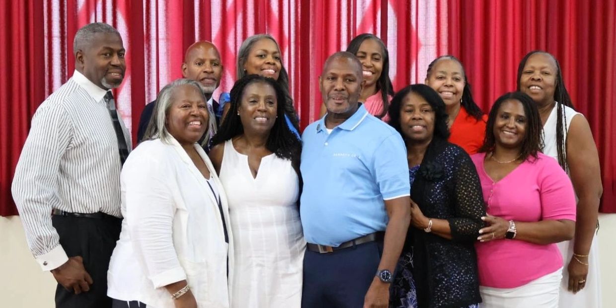 A group of twelve adults posing indoors in front of red curtains, smiling.