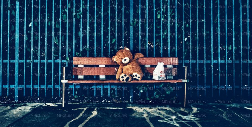 Teddy bear sitting alone on a wooden bench by a blue fence at night. Photography prints, wall art and fine art prints are available at our print store.