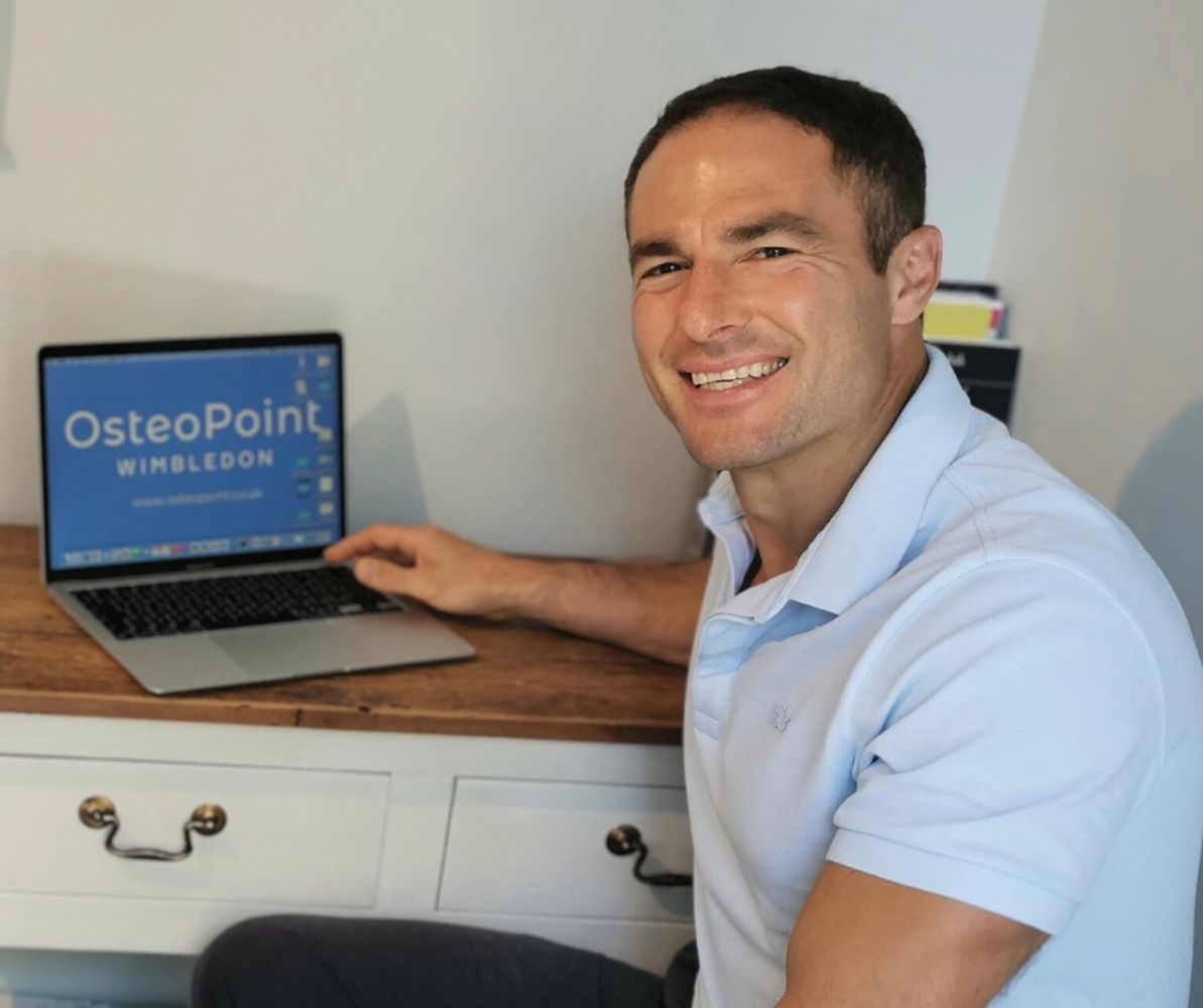 Smiling osteopath with brown hair sitting at a desk in clinic room, wearing a white polo shirt