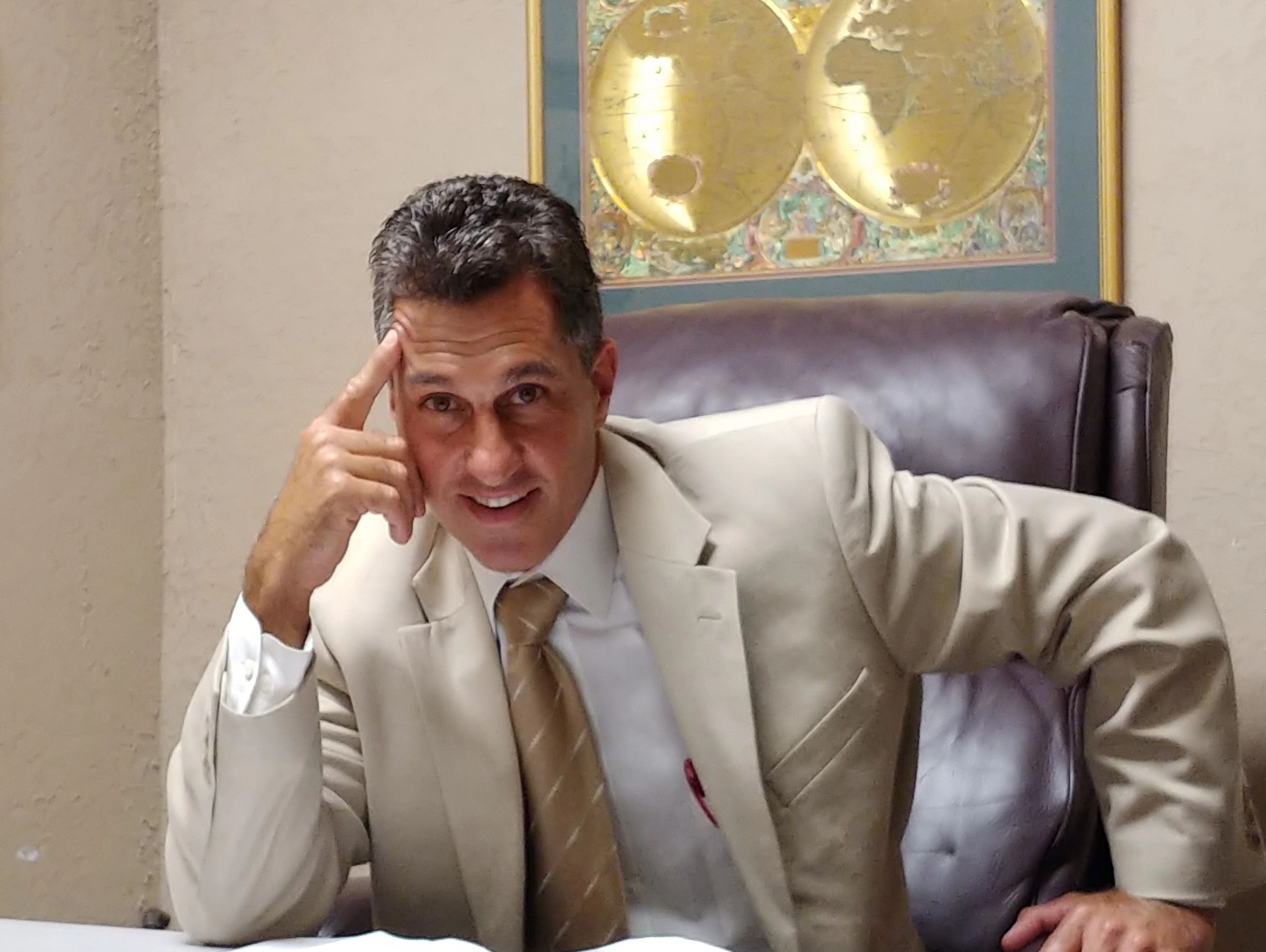 Man in beige suit sitting at desk, smiling thoughtfully.
